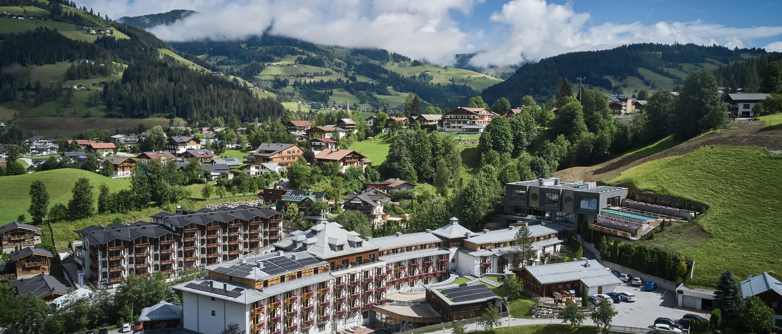 Luftaufnahme von Sporthotel Wagrain vor grüner Hügellandschaft unter blauem Himmel mit Wolken