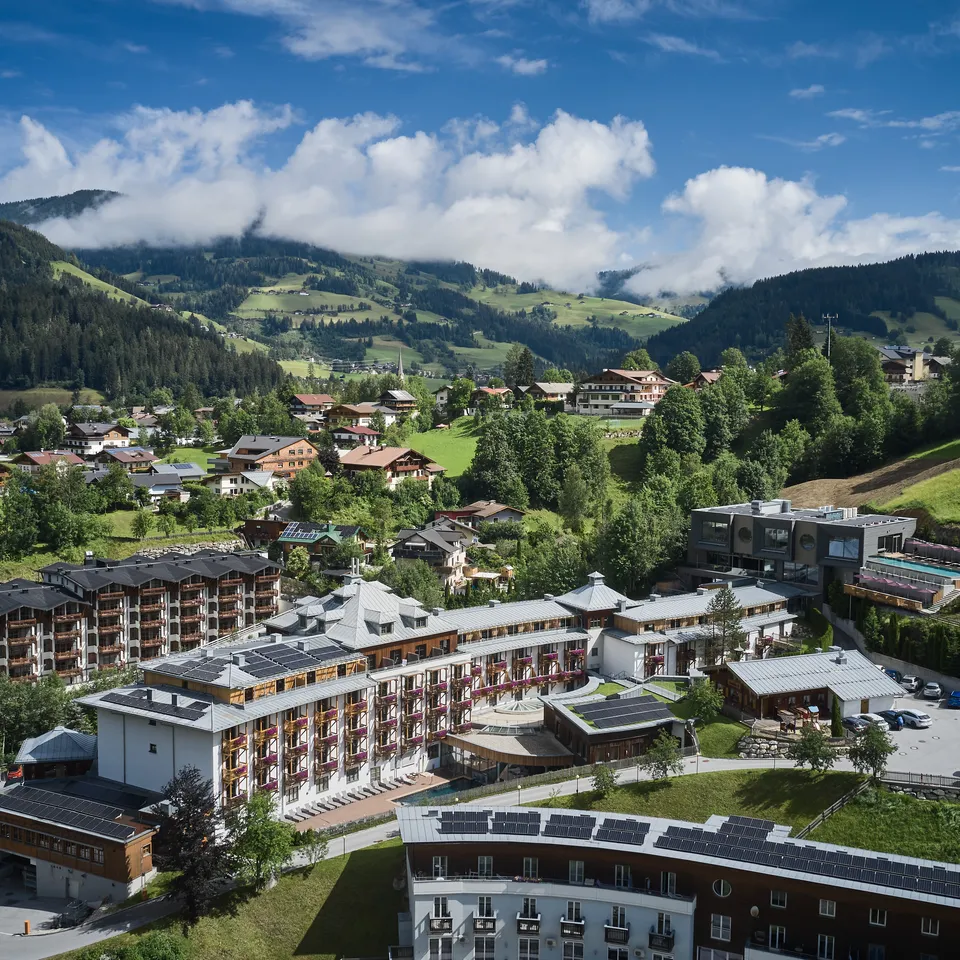 Luftaufnahme von Sporthotel Wagrain vor grüner Hügellandschaft unter blauem Himmel mit Wolken