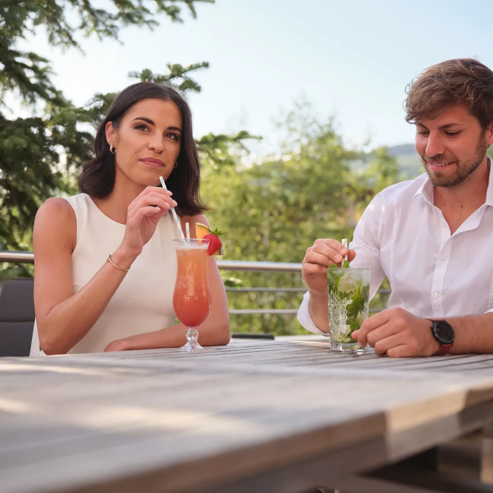 Frau trinkt Cocktail mit Strohhalm, Mann rührt Drink mit Minze auf Terrasse