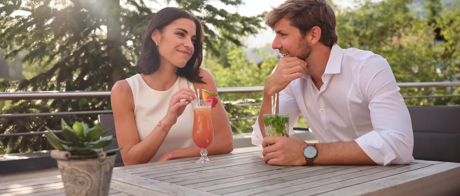 Frau und Mann sitzen mit Cocktails an Holztisch im Freien