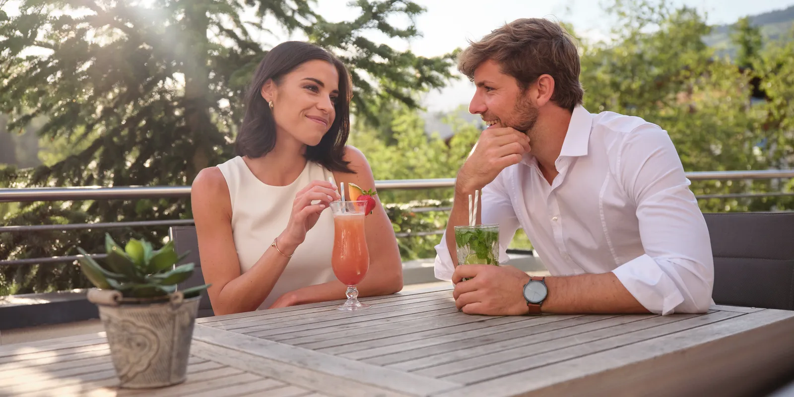 Frau und Mann sitzen mit Cocktails an Holztisch im Freien