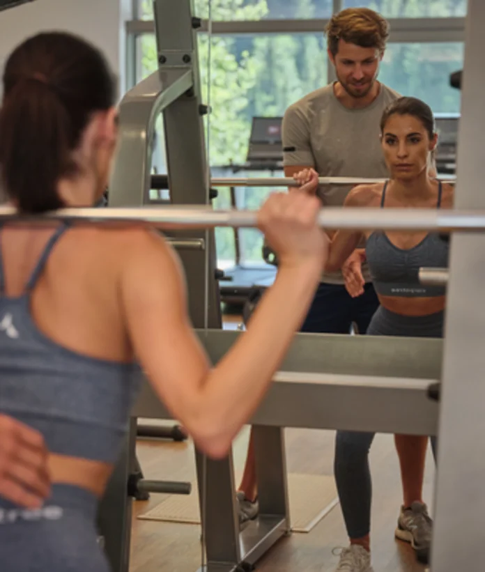 Frau trainiert mit Langhantel im Fitnessstudio, Trainer gibt Hilfestellung