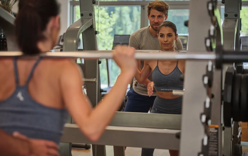 Frau trainiert mit Langhantel im Fitnessstudio, Mann gibt Hilfestellung