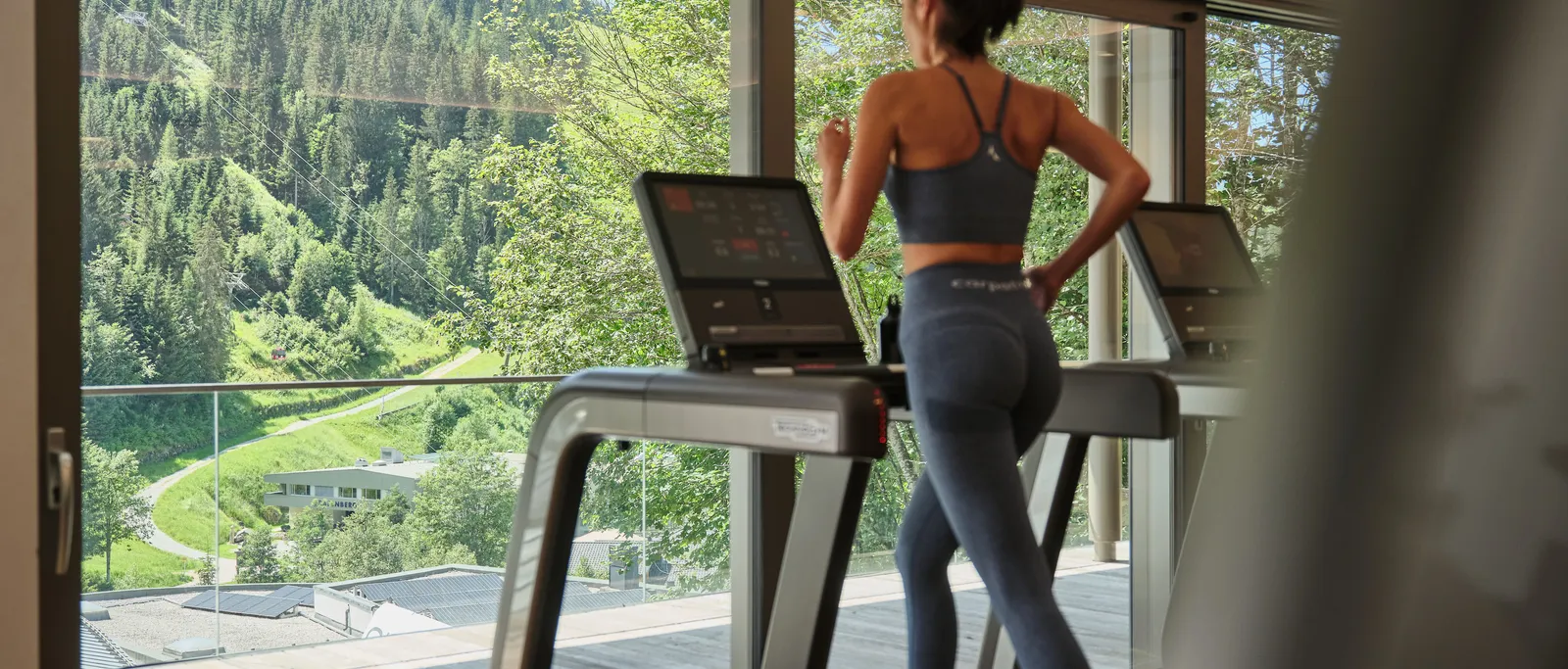 Woman in sportswear running on treadmill in front of panoramic window with mountain view