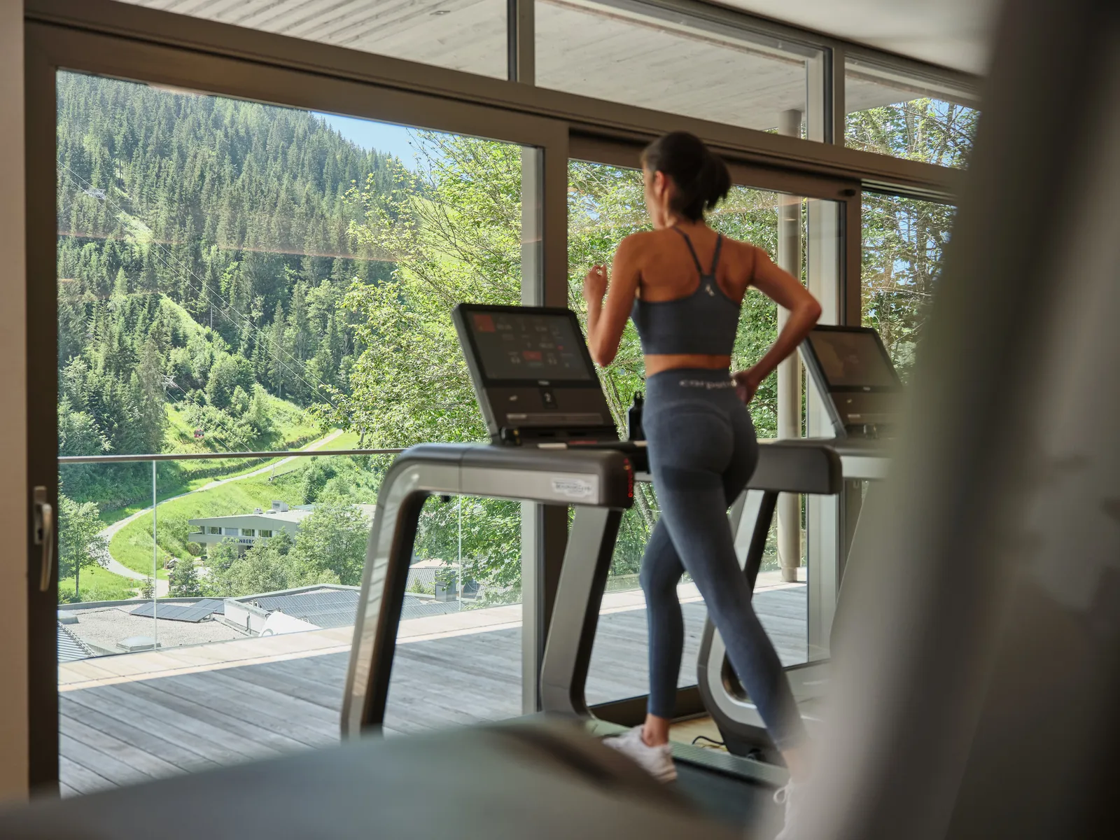 Frau in Sportkleidung läuft auf Laufband vor Panoramafenster mit Bergblick