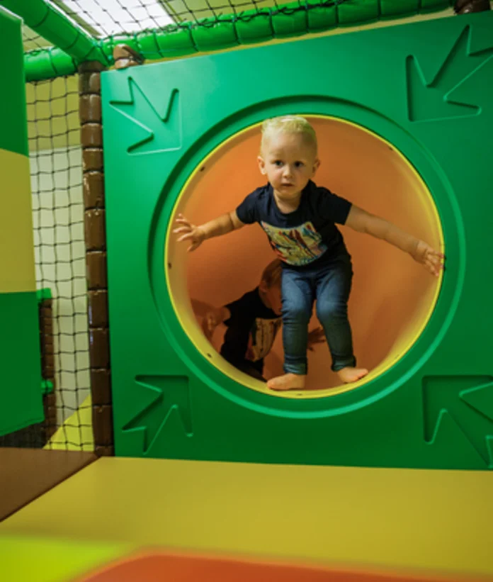 Kind klettert durch grüne Röhre im Indoor-Spielbereich mit Netzen und Polstern