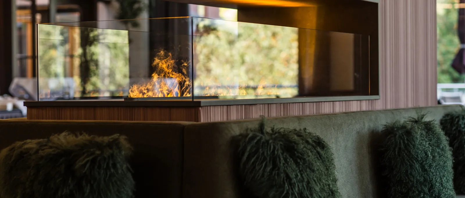 Modern fireplace with glass front and flames above sofa with decorative cushions in front of wooden wall