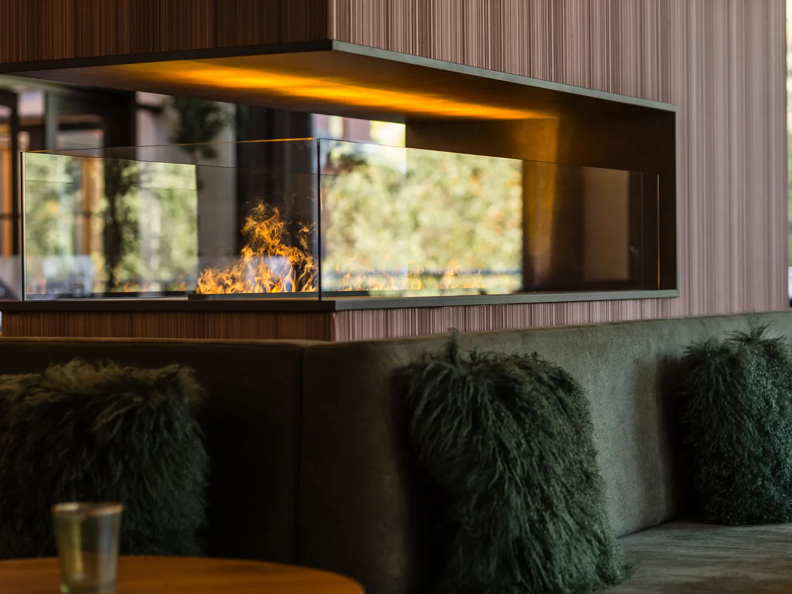 Moderner Kamin mit Glasfront und Flammen über Sofa mit Zierkissen vor Holzwand