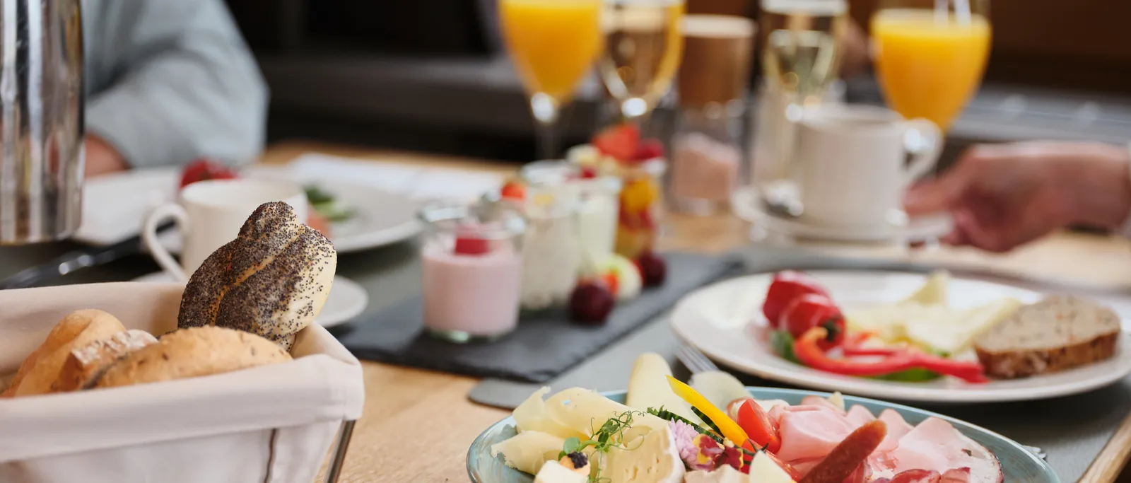 Reichhaltiges Frühstücksbuffet mit Wurst, Käse, Brötchen, Joghurt und Orangensaft auf einem Holztisch