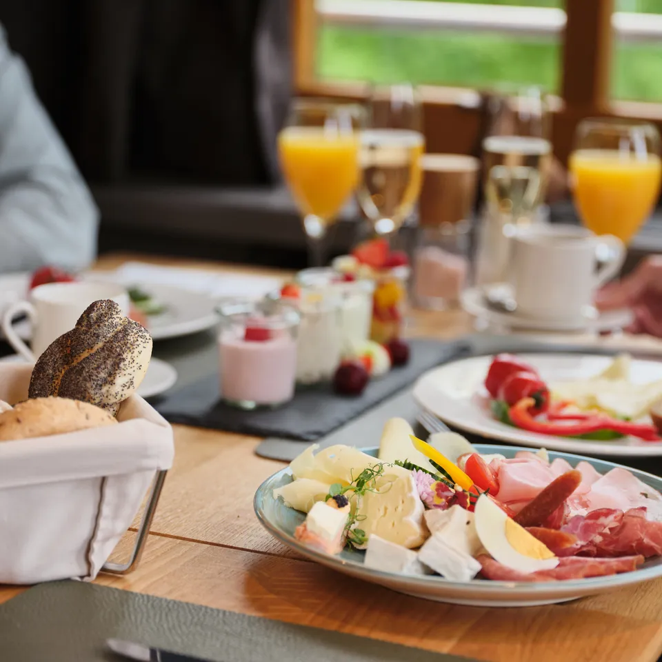 Reichhaltiges Frühstücksbuffet mit Wurst, Käse, Brötchen, Joghurt und Orangensaft auf einem Holztisch