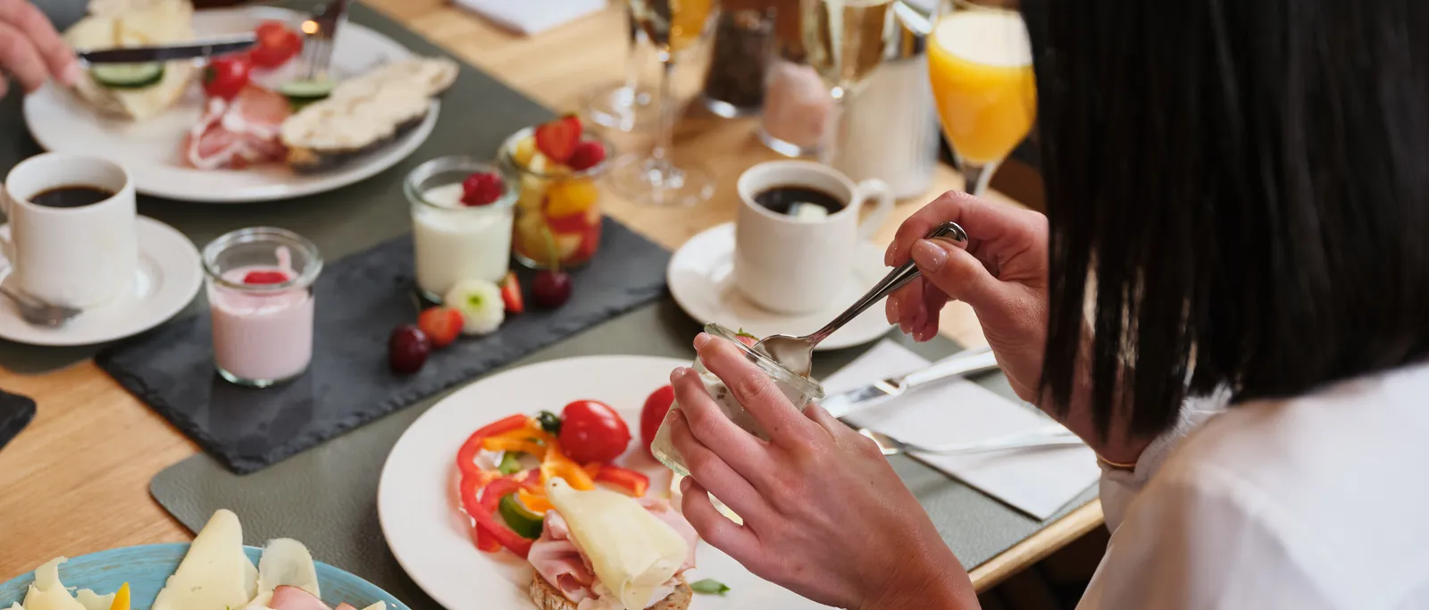 Person eating sandwich at breakfast table with sausage, cheese, yogurt, fruit and drinks