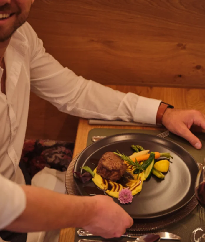 Laughing man presents plate with meat, vegetables and edible flower next to wine glass