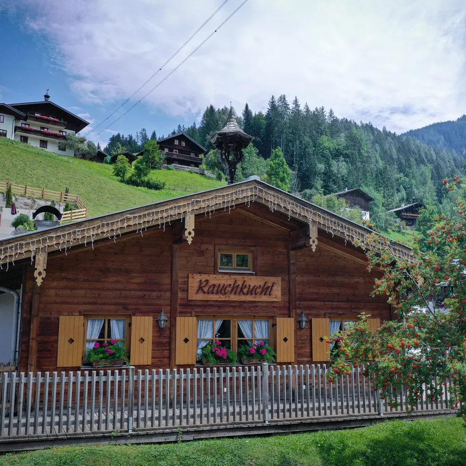 Braunes Holzhaus "Rauchkuchl" mit gelben Fensterläden vor grüner Hügellandschaft