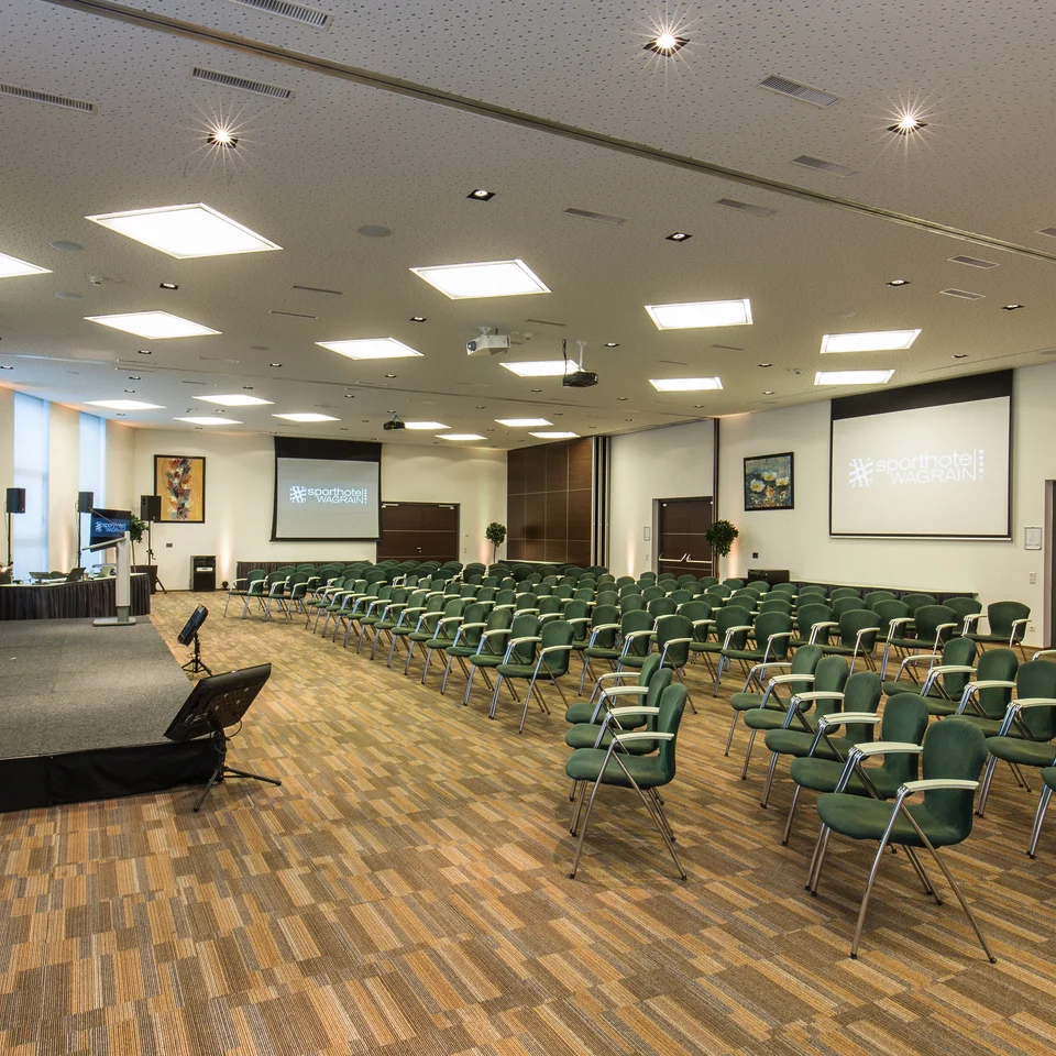 Seminarraum mit Bühne, Leinwänden und grünen Stühlen im Sporthotel Wagrain