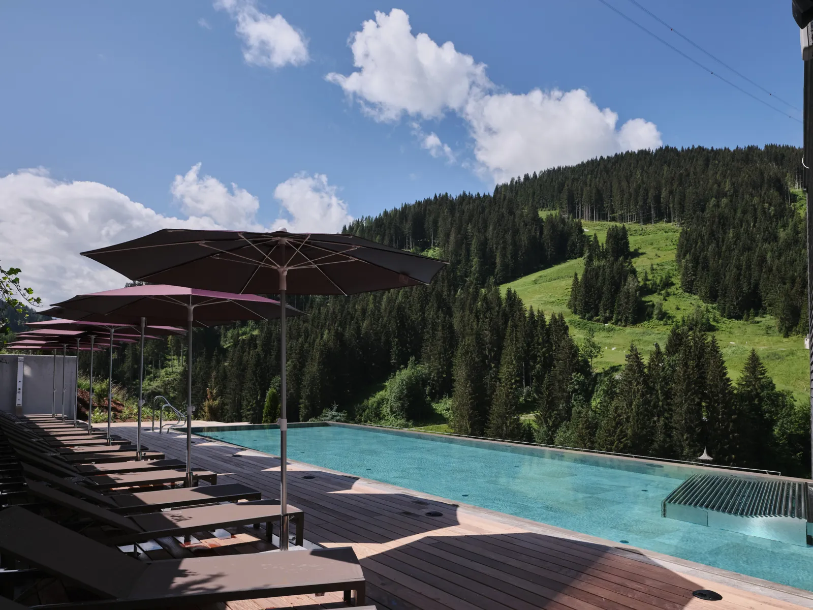 Pool with loungers in front of a wooded mountain slope under a blue sky