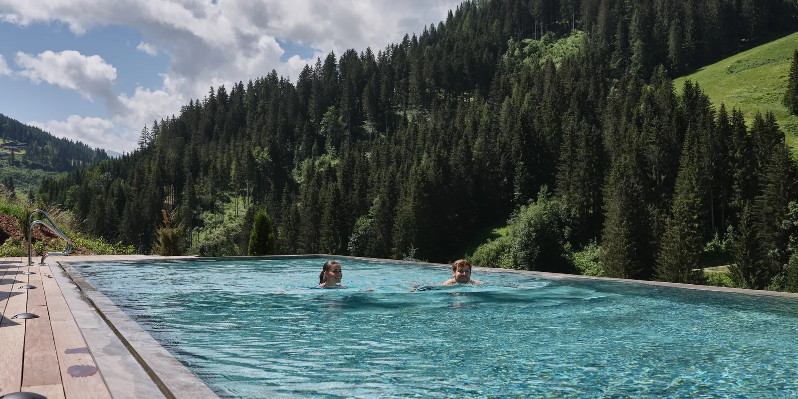 Zwei Personen schwimmen in einem Pool vor bewaldeten Bergen unter einem bewölkten Himmel