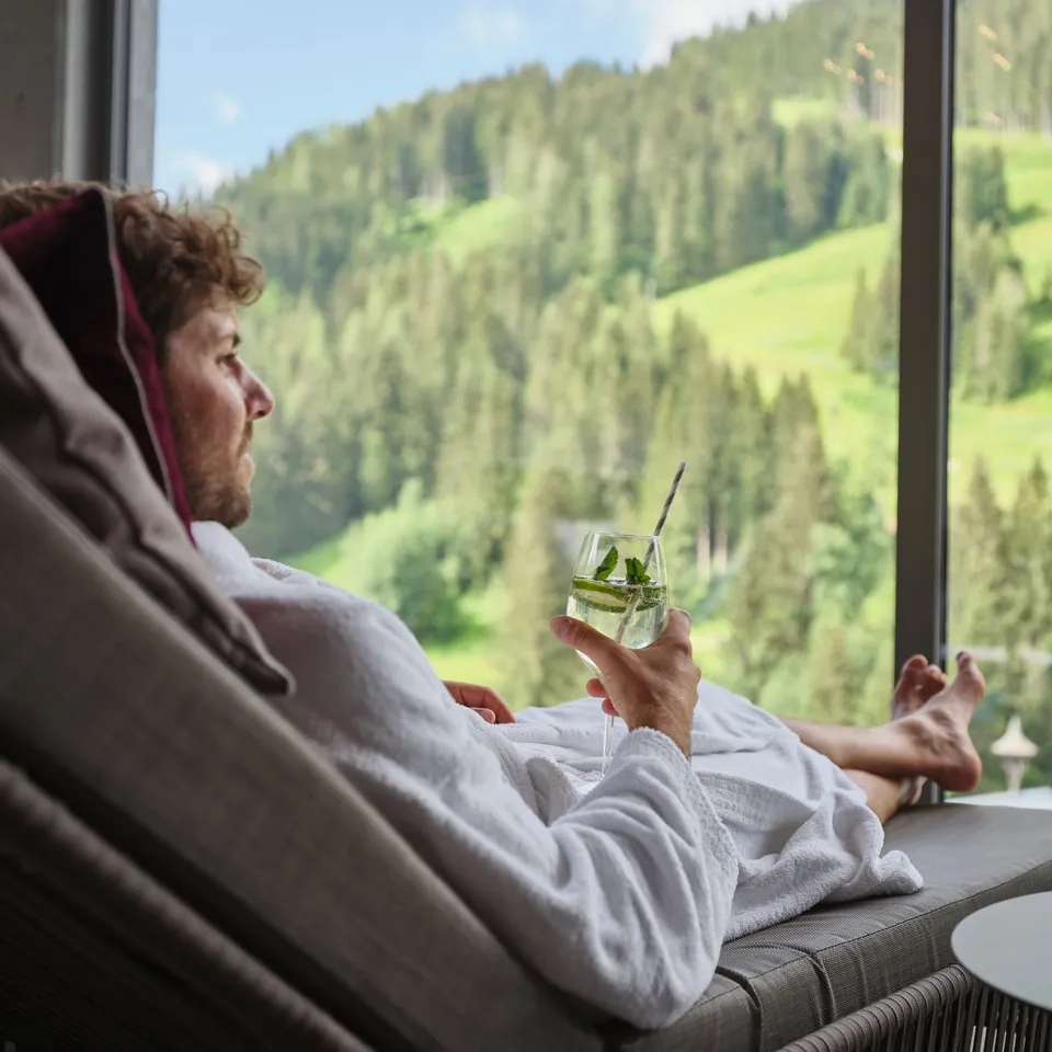 Mann im Bademantel entspannt mit Drink vor Fenster mit Bergblick