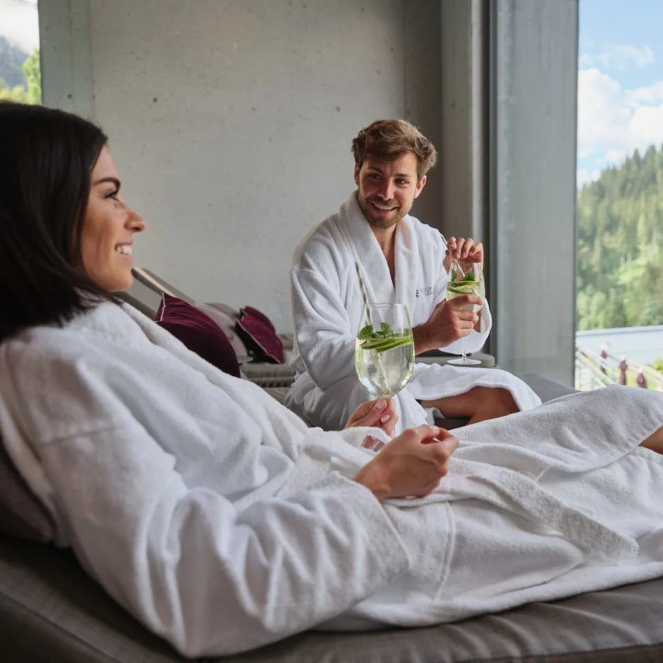 Paar in Bademänteln entspannt mit Getränken vor Fenster mit Bergblick
