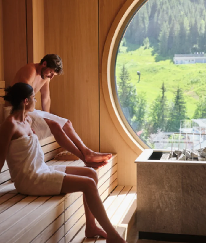 Couple in towels sitting in sauna in front of round window overlooking green landscape
