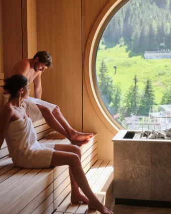 Couple in towels sitting in sauna in front of round window overlooking green landscape