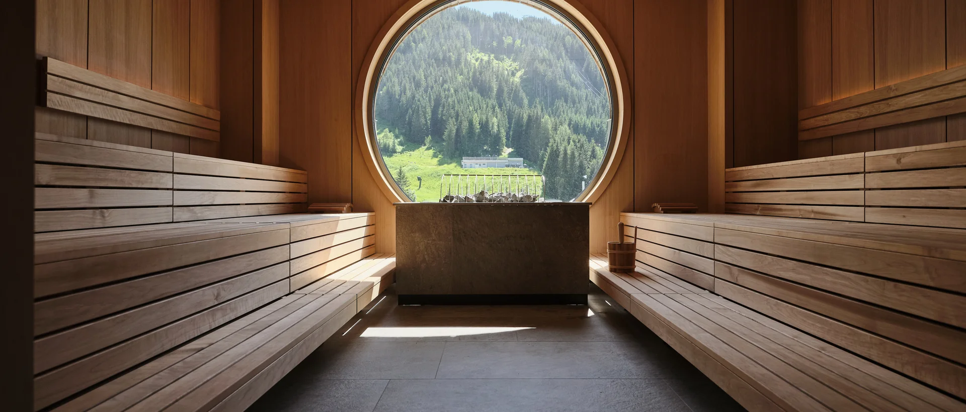Holzsauna mit runden Fensterblick auf grüne Berglandschaft