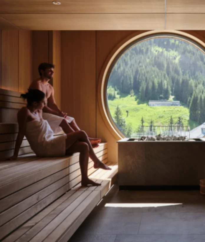 Sauna mit zwei Personen vor rundem Fenster mit Blick auf grüne Landschaft
