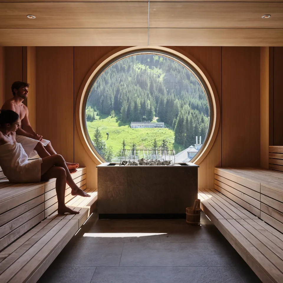 Sauna mit zwei Personen vor rundem Fenster mit Blick auf grüne Landschaft