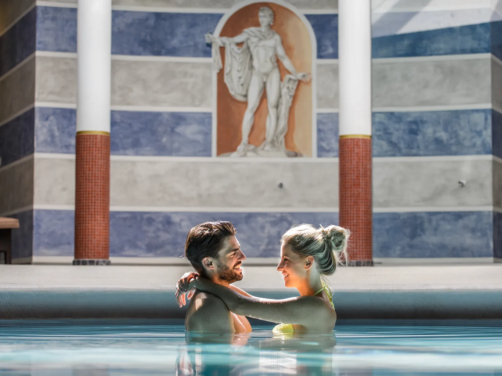 Couple in the outdoor pool of the Sporthotel Wagrain in front of a mural with a statue
