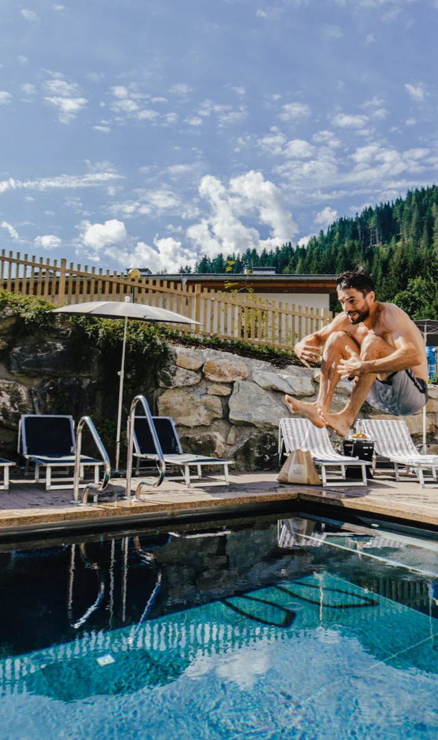 Mann springt von Liege in Außenpool vor Bergkulisse