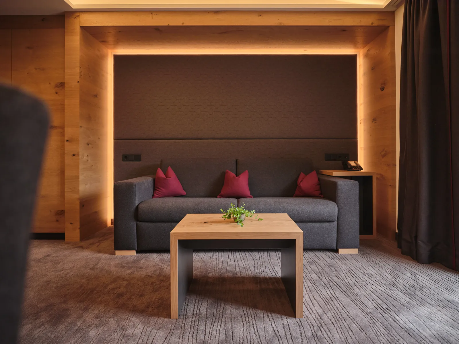 Graues Sofa mit roten Kissen und Holztisch vor dunkler Wand mit indirekter Beleuchtung