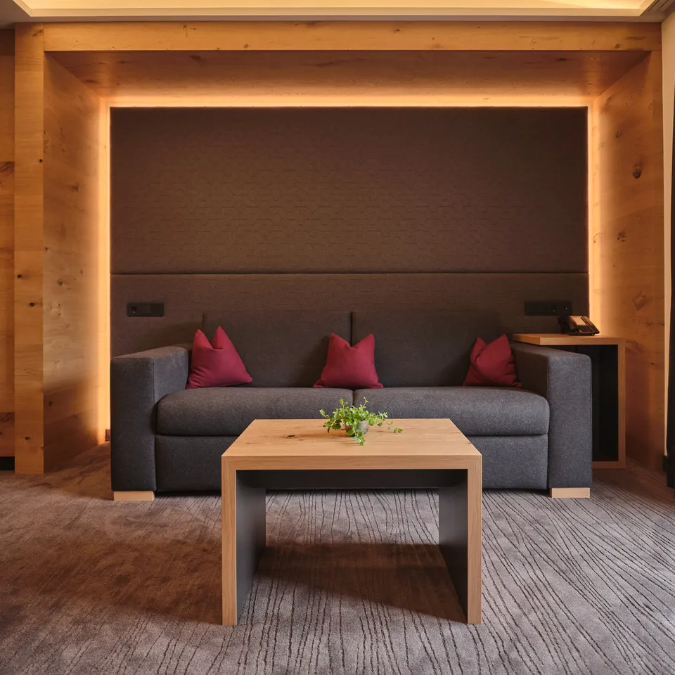 Graues Sofa mit roten Kissen und Holztisch vor dunkler Wand mit indirekter Beleuchtung