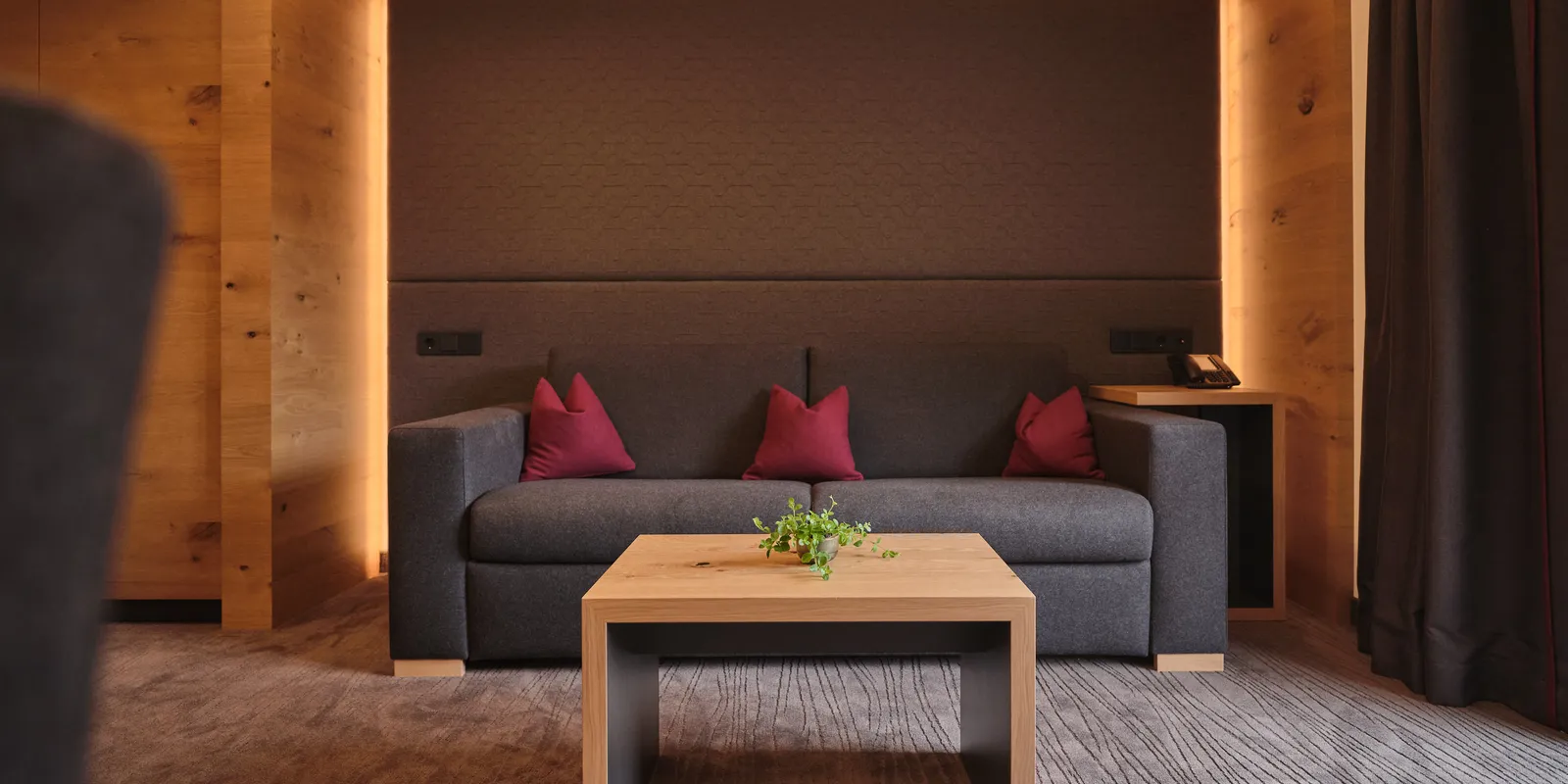 Graues Sofa mit roten Kissen und Holztisch vor dunkler Wand mit indirekter Beleuchtung