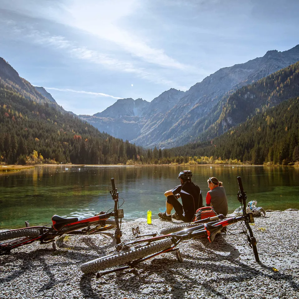Zwei Personen sitzen am See mit Fahrrädern, im Hintergrund Berge und Wald