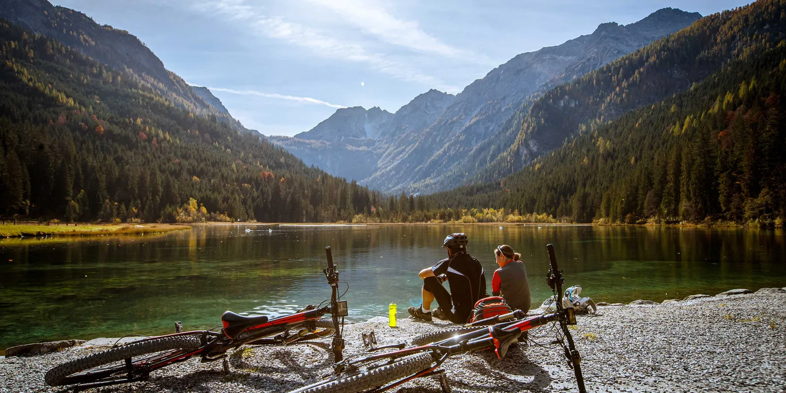 Two people sitting by the lake with bicycles, mountains and forest in the background