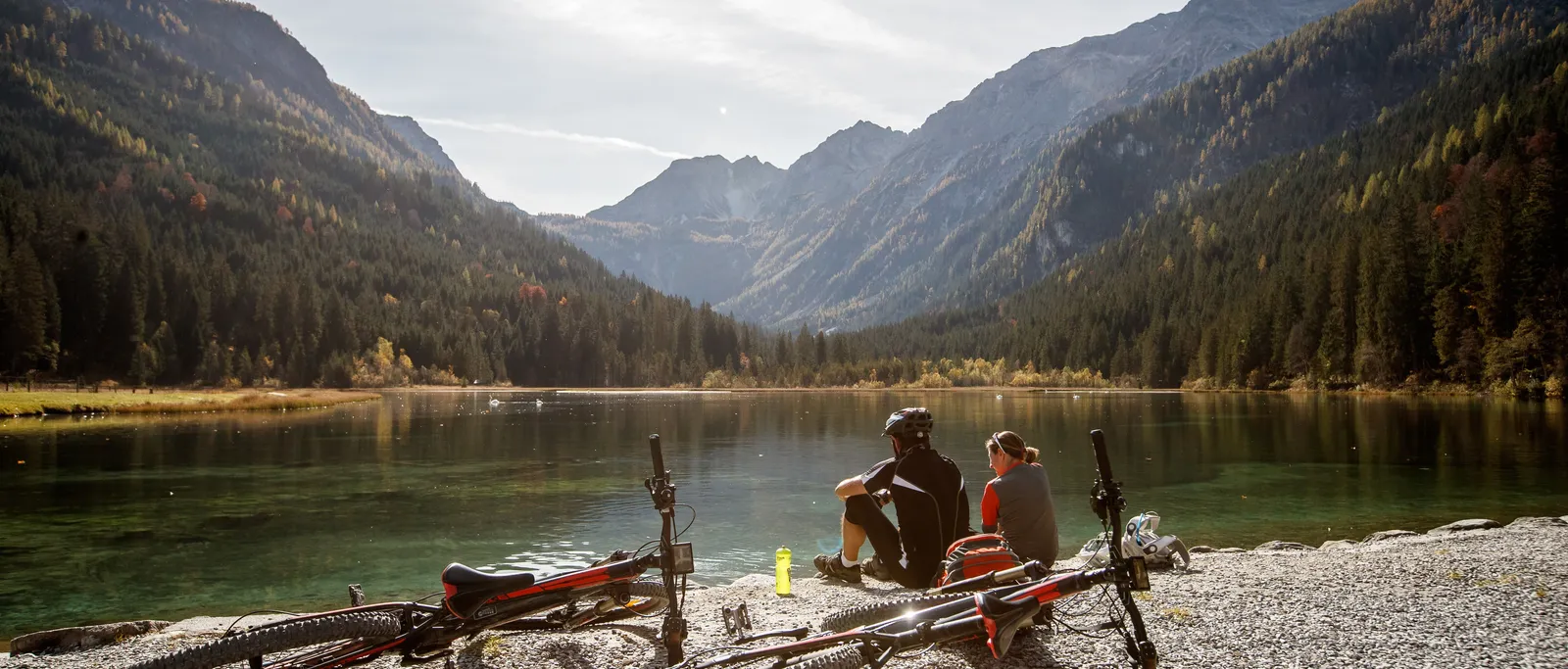 Radfahrerpaar sitzt am See mit Fahrrädern im Vordergrund und Bergen im Hintergrund