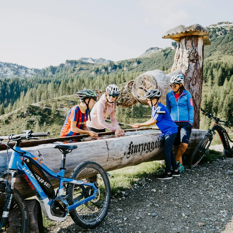Familie macht E-Bike-Tour und erfrischt sich an einem Brunnen vor Bergkulisse