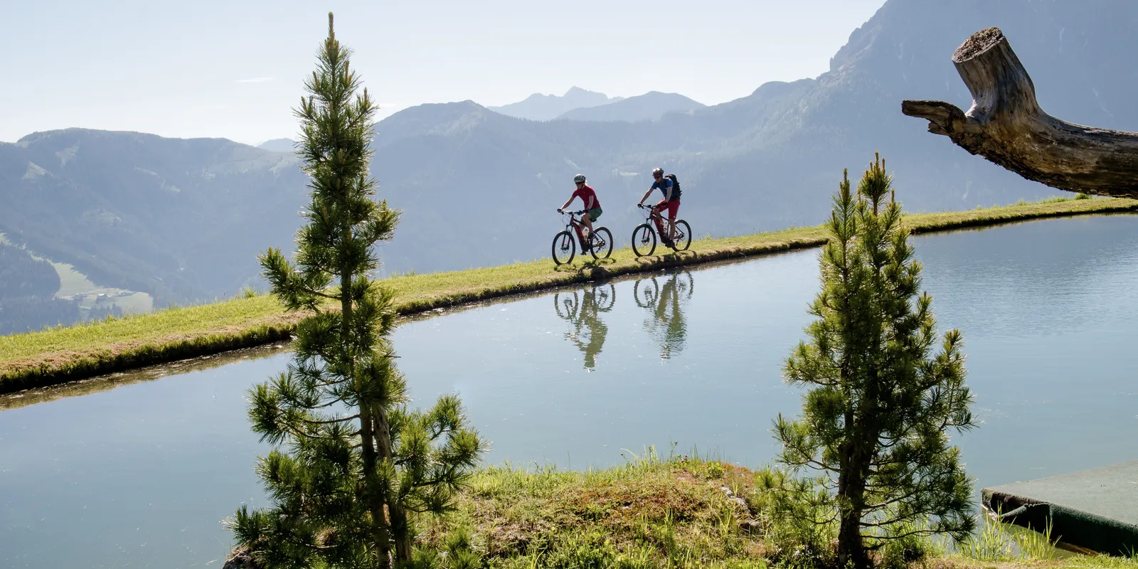Two e-mountain bikers ride along the lake shore with reflections in the water in front of a mountain backdrop