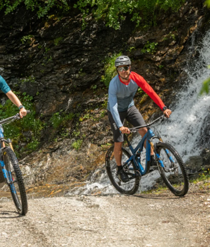 Two mountain bikers ride on a gravel road in front of a waterfall