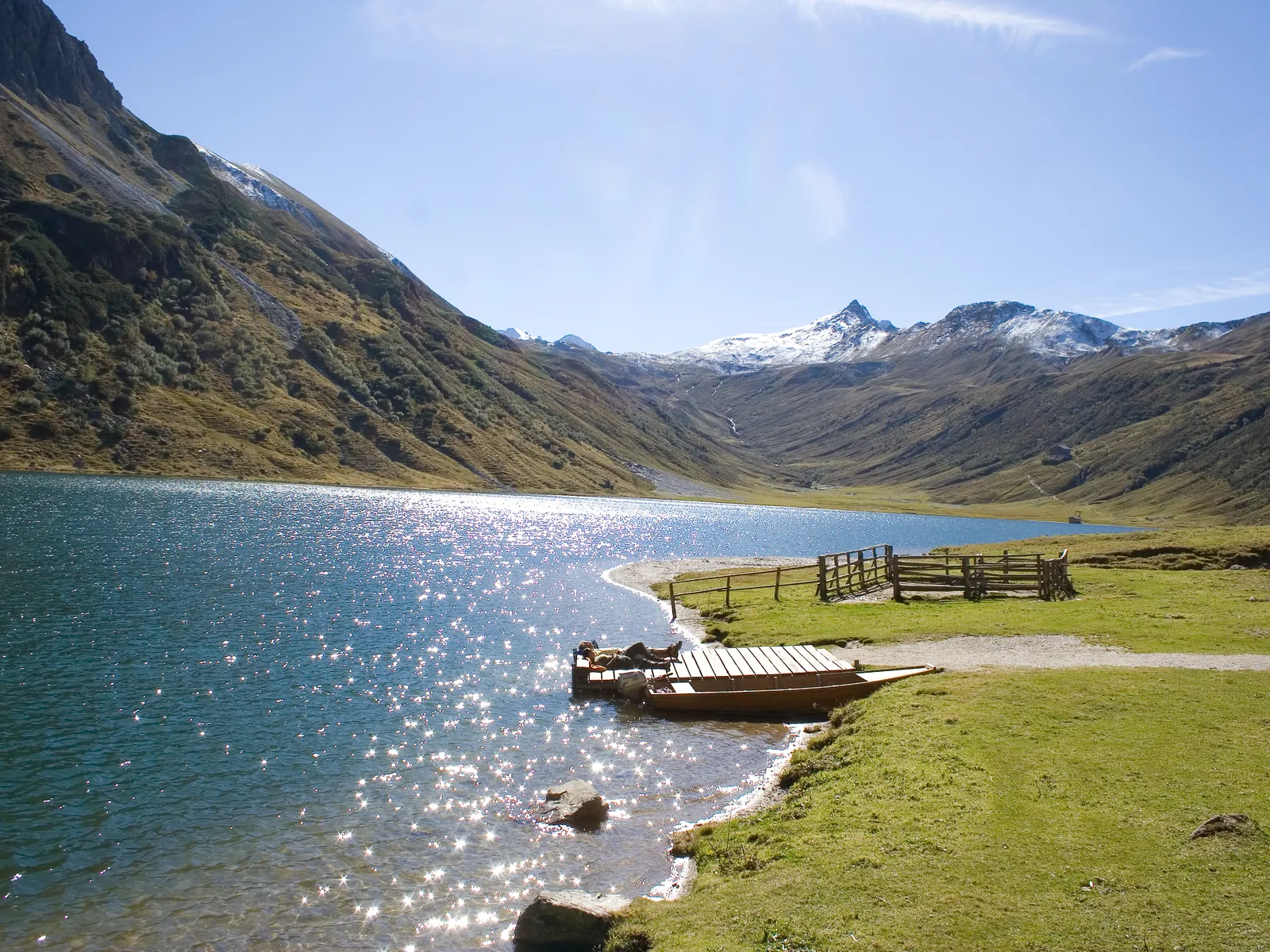 Tappenkarsee mit Steg, umgeben von grünen Wiesen und Bergen unter blauem Himmel
