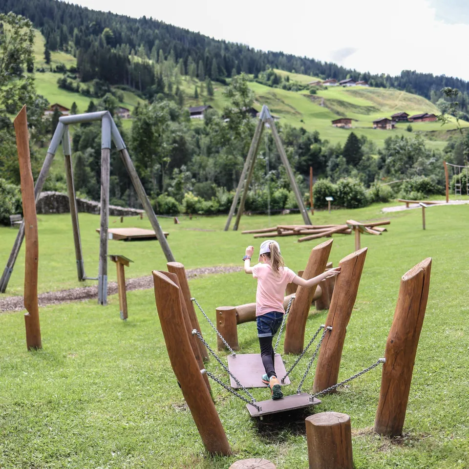 Mädchen balanciert auf Spielplatz mit Holzelementen vor grüner Hügellandschaft