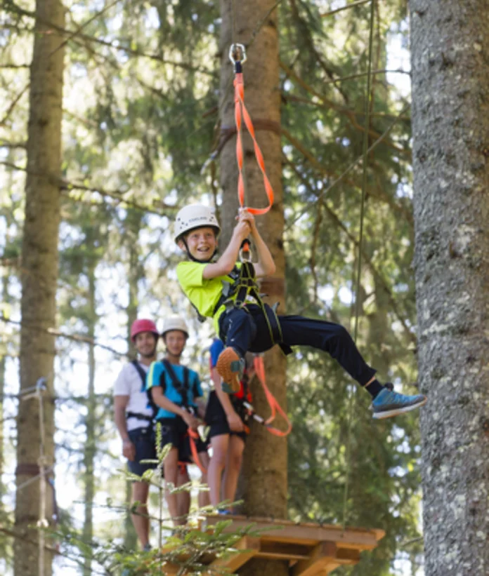Kind mit Helm fährt Seilrutsche im Kletterpark, andere Personen schauen zu