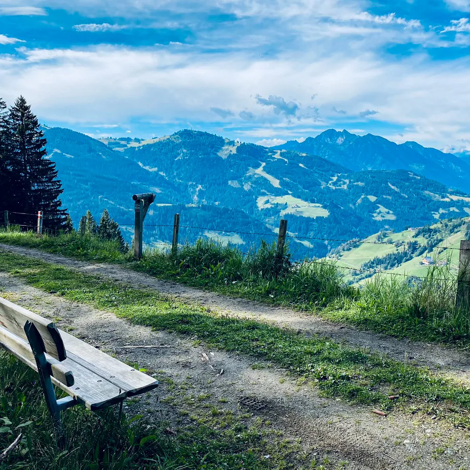 Berglandschaft mit Weg, Bank und bewaldeten Hügeln unter blauem Himmel