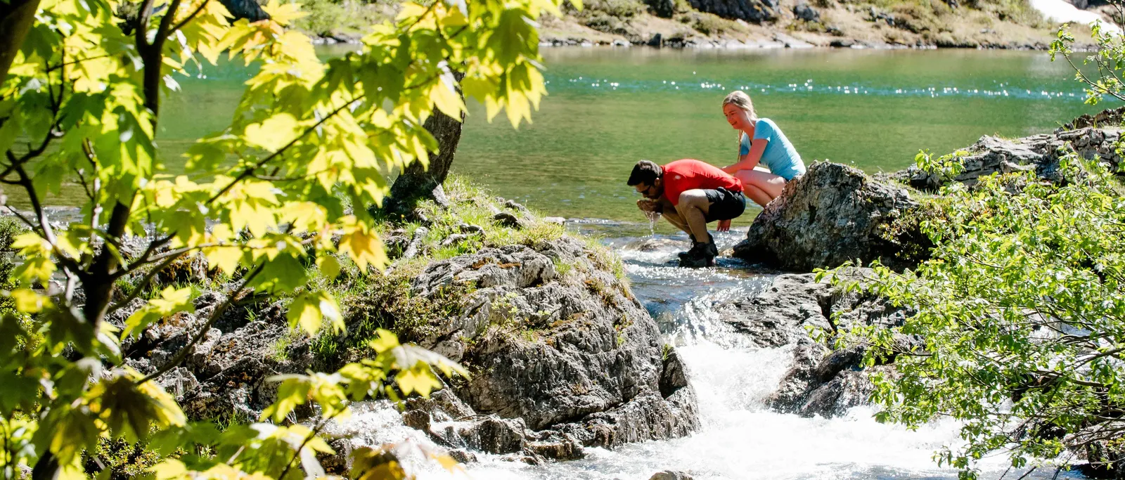 Mann beugt sich an Fluss, Frau sitzt auf Felsen, Bäume im Vordergrund
