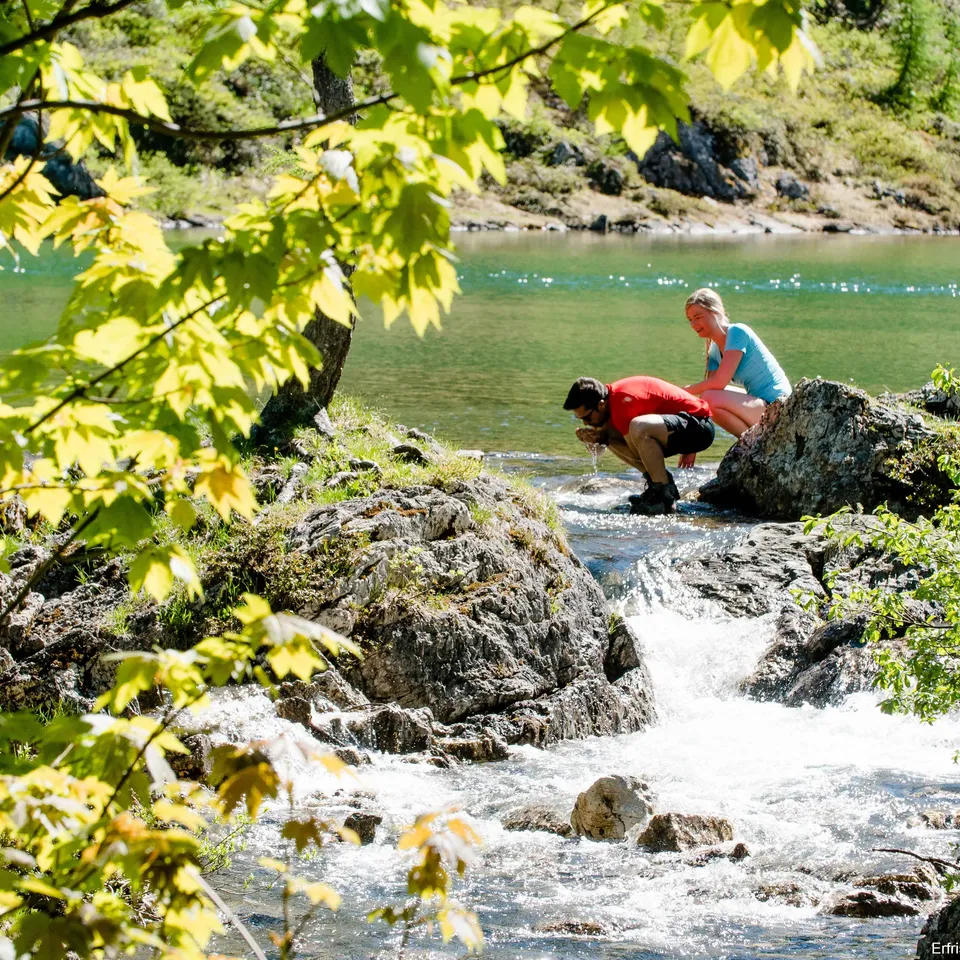 Mann beugt sich an Fluss, Frau sitzt auf Felsen, Bäume im Vordergrund