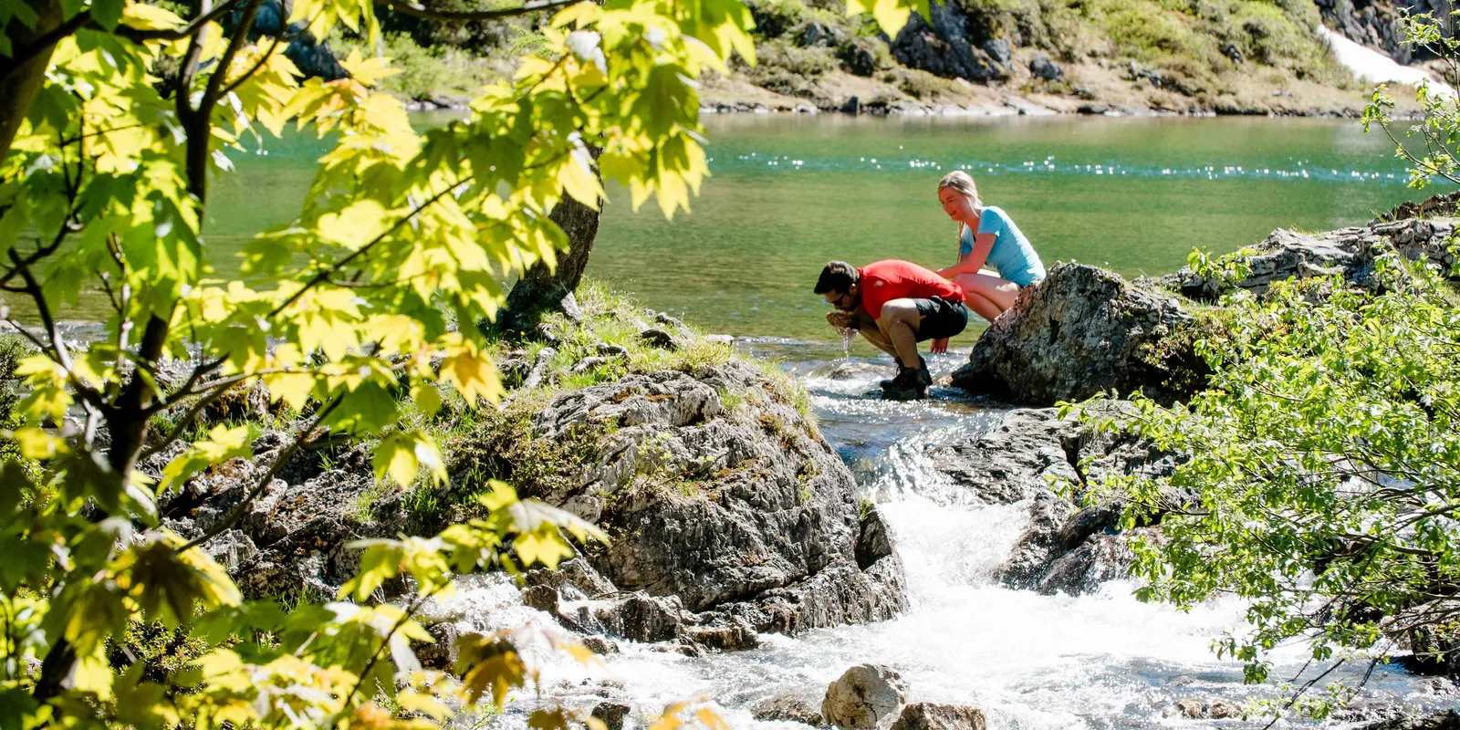 Mann beugt sich an Fluss, Frau sitzt auf Felsen, Bäume im Vordergrund