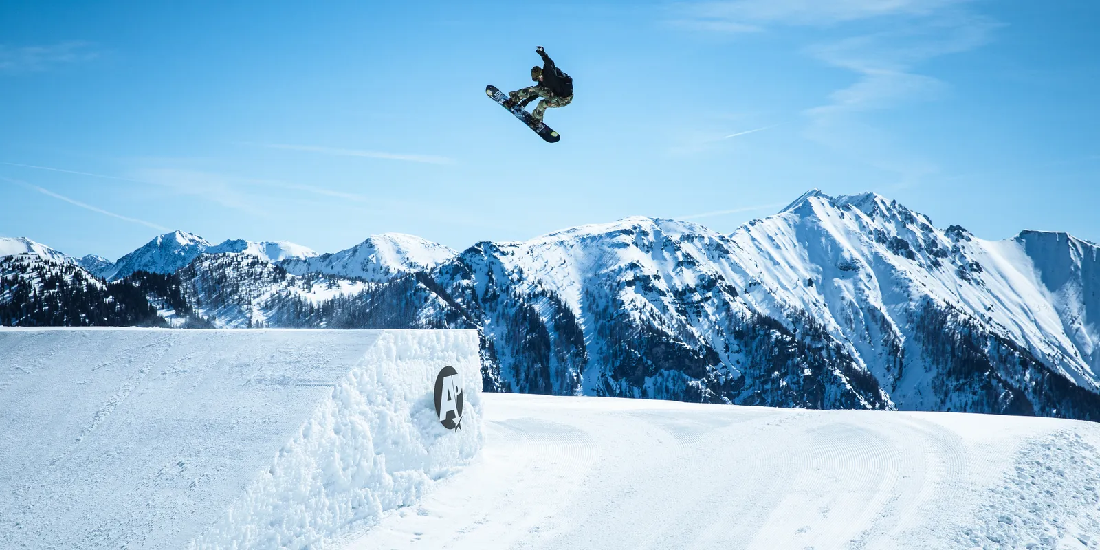 Snowboarder springt über Schanze vor schneebedeckten Bergen unter blauem Himmel