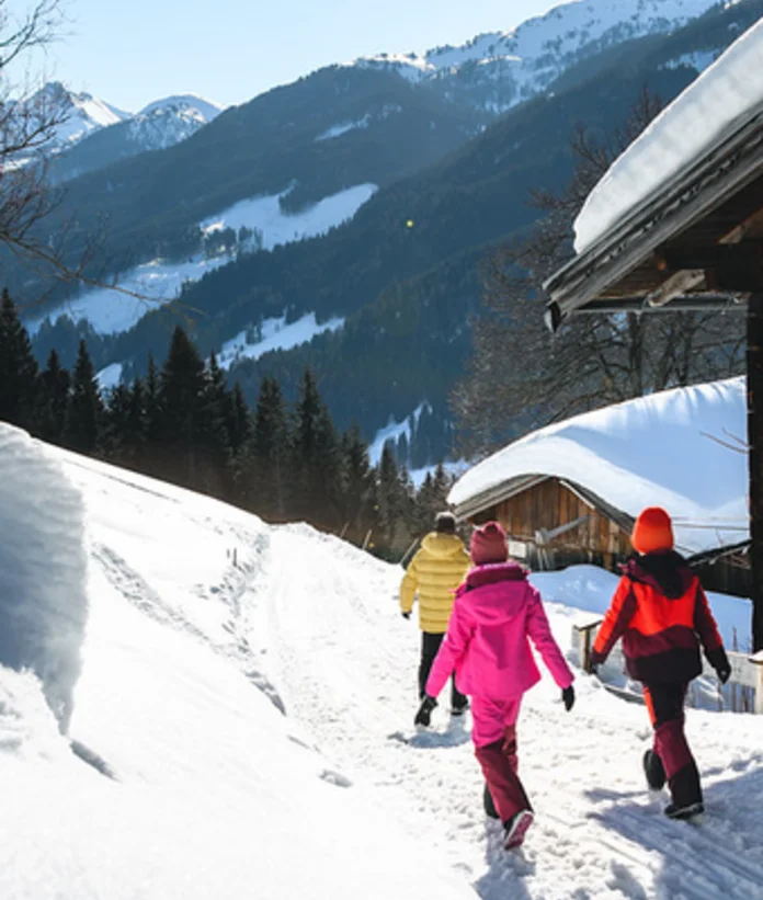 Winterwanderung: Kinder laufen auf verschneitem Pfad an Almen vorbei, im Hintergrund Berge