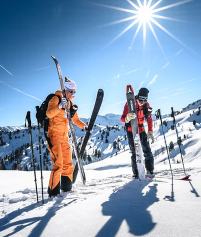 Zwei Skitourengeher mit Skiern und Stöcken im Schnee vor Bergpanorama unter strahlender Sonne
