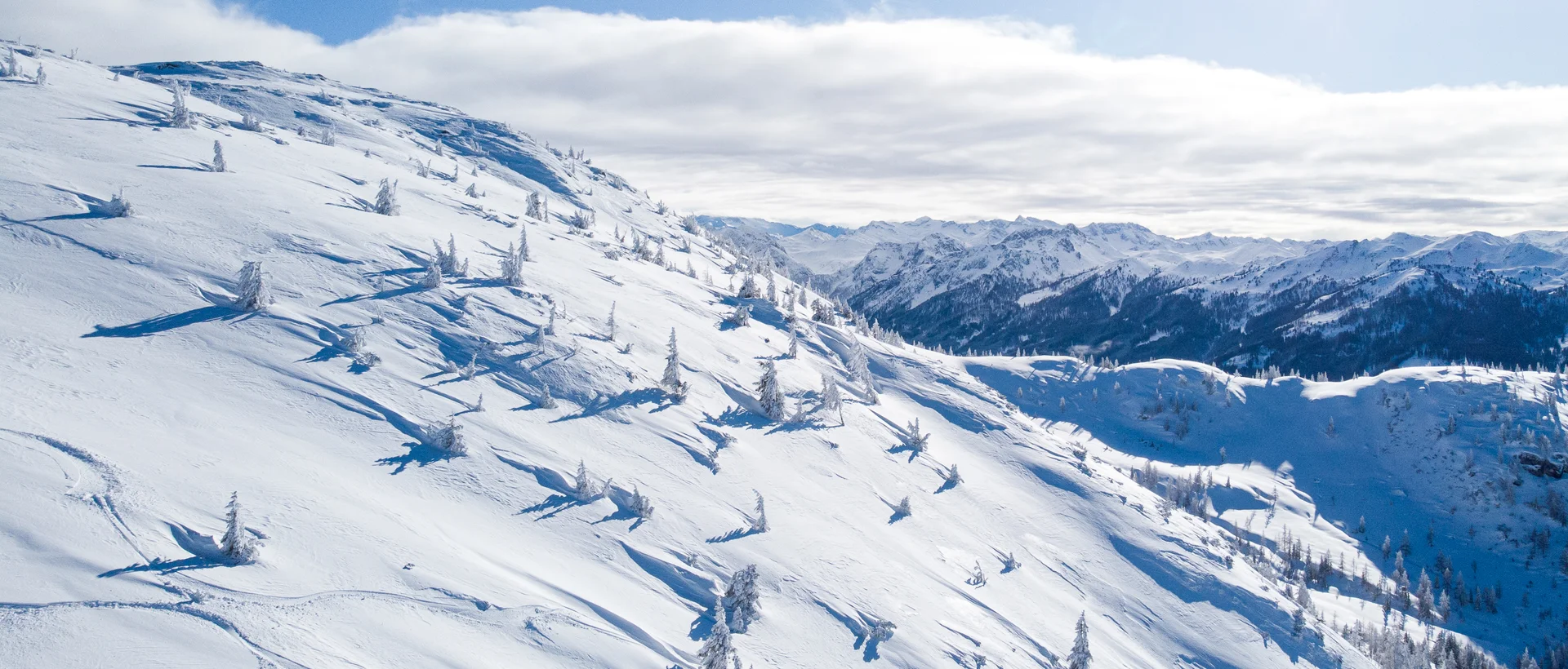 Verschneiter Berghang mit Bäumen unter blauem Himmel mit Wolken