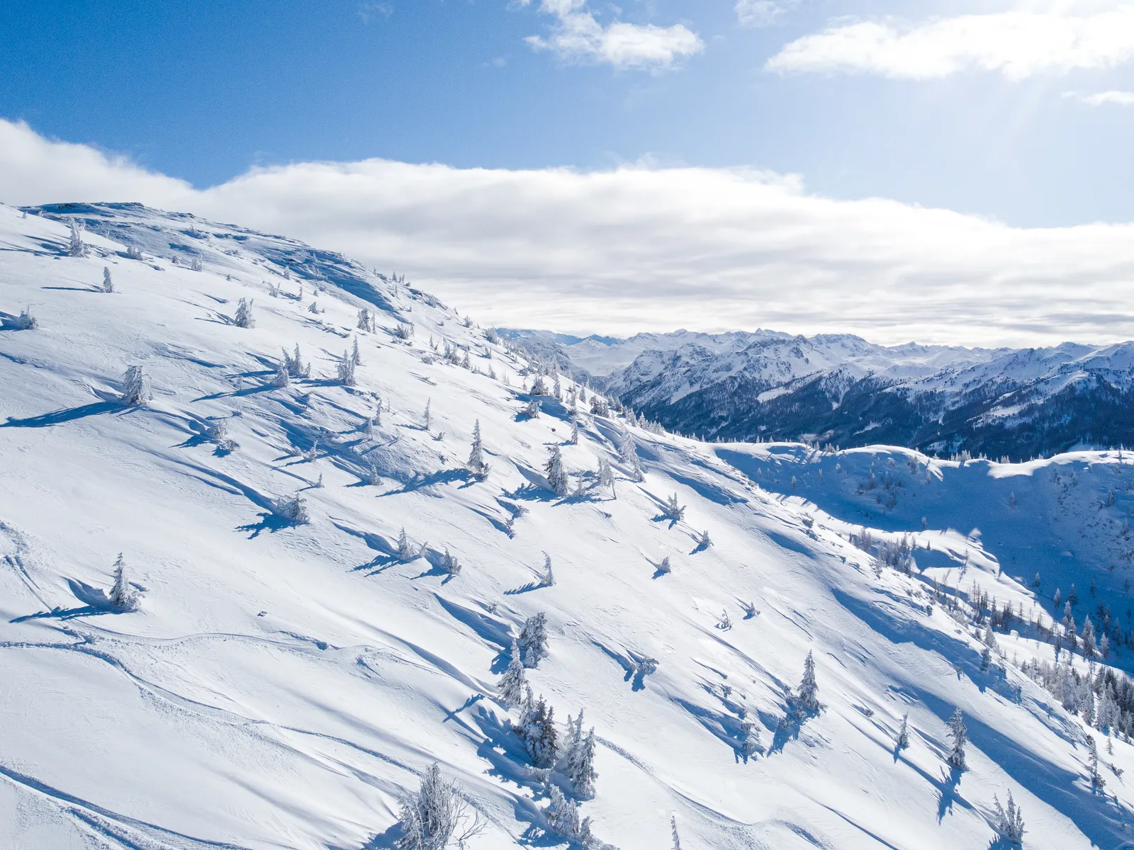 Verschneiter Berghang mit Bäumen unter blauem Himmel mit Wolken